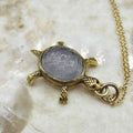 A vintage 18k gold turtle charm with a carved amethyst shell and red stone eyes, displayed on a gold-filled chain against a marble background.