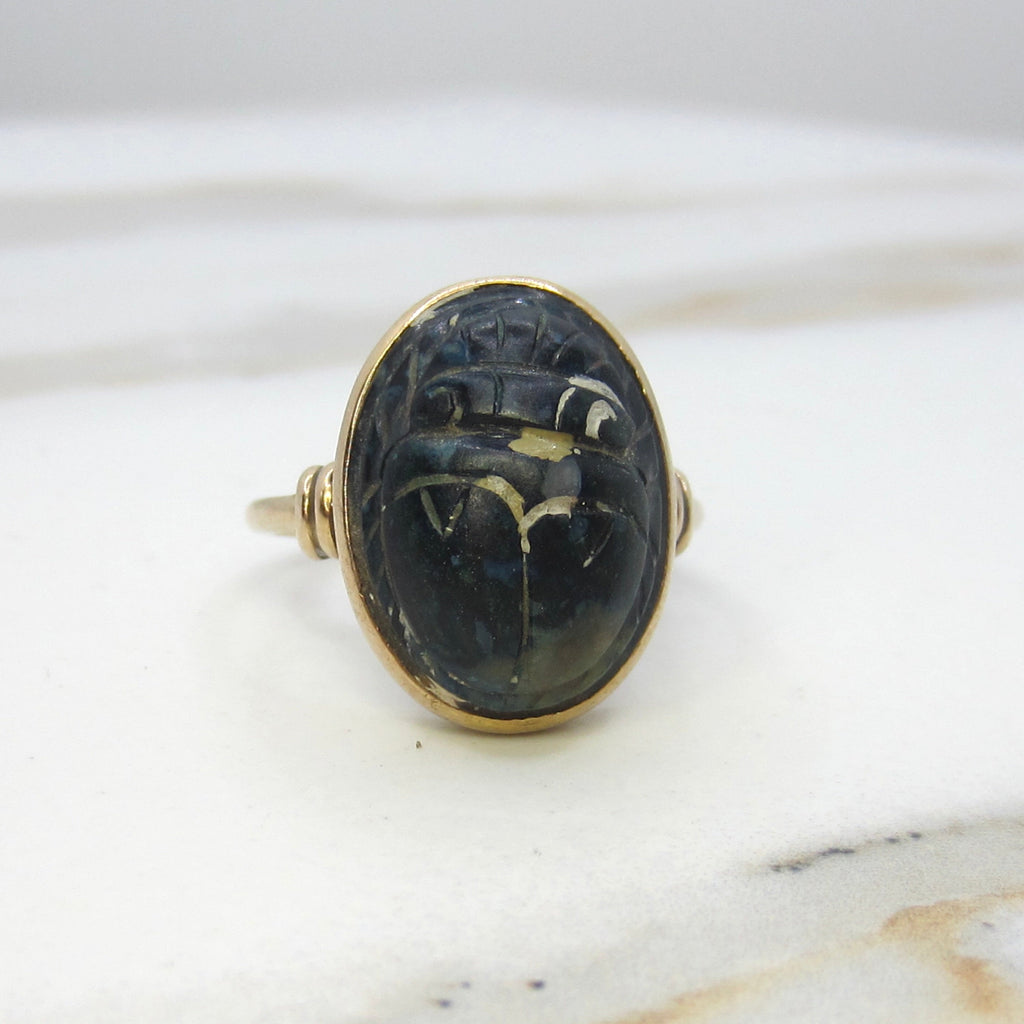 A vintage gold ring featuring an oval black cabochon stone carved with ancient hieroglyphic symbols, resting on a white marble surface.