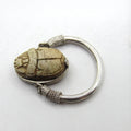 A silver-toned ring featuring a carved beetle pendant with a textured, weathered beige-brown finish, set against a plain white background.