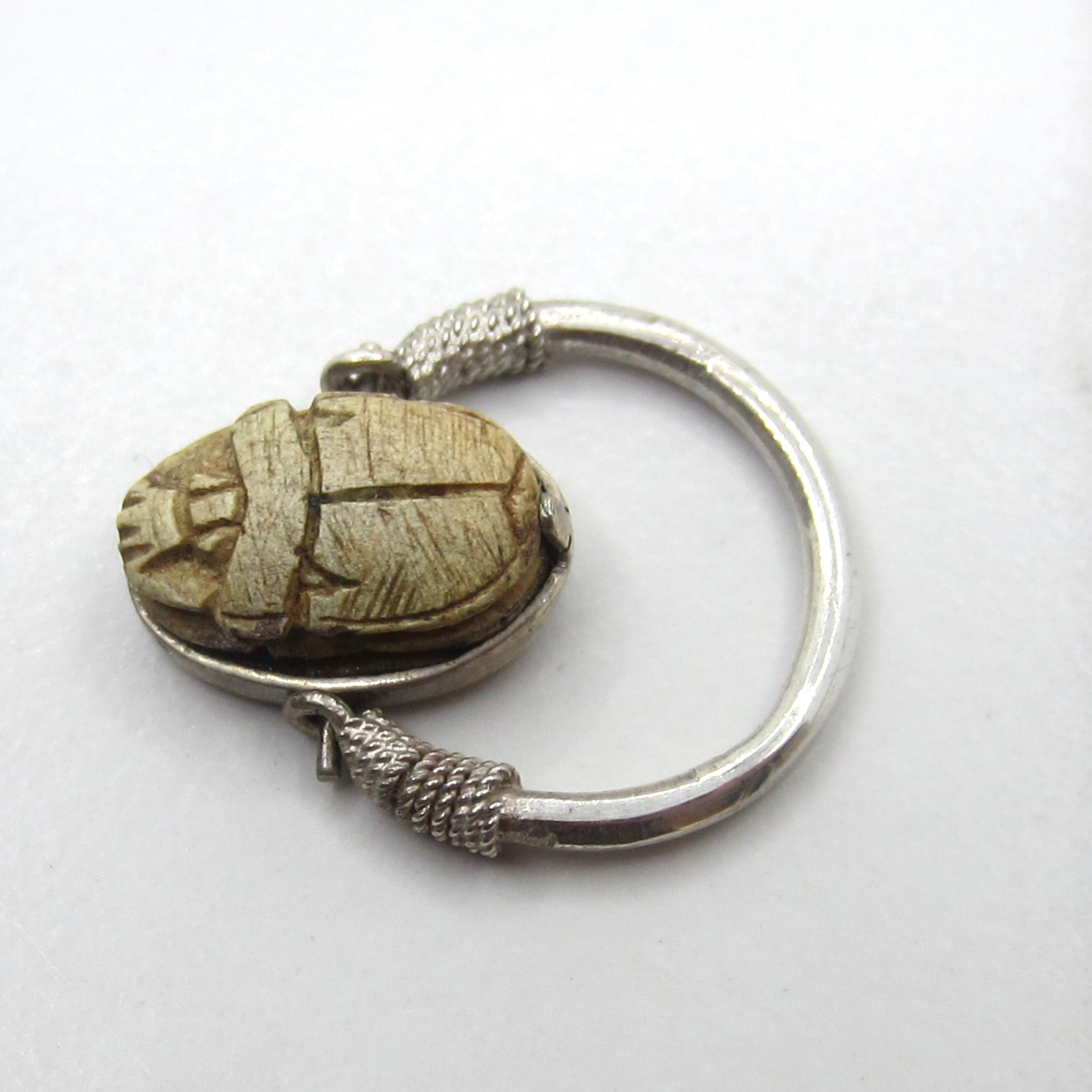 A silver-toned ring featuring a carved beetle pendant with a textured, weathered beige-brown finish, set against a plain white background.