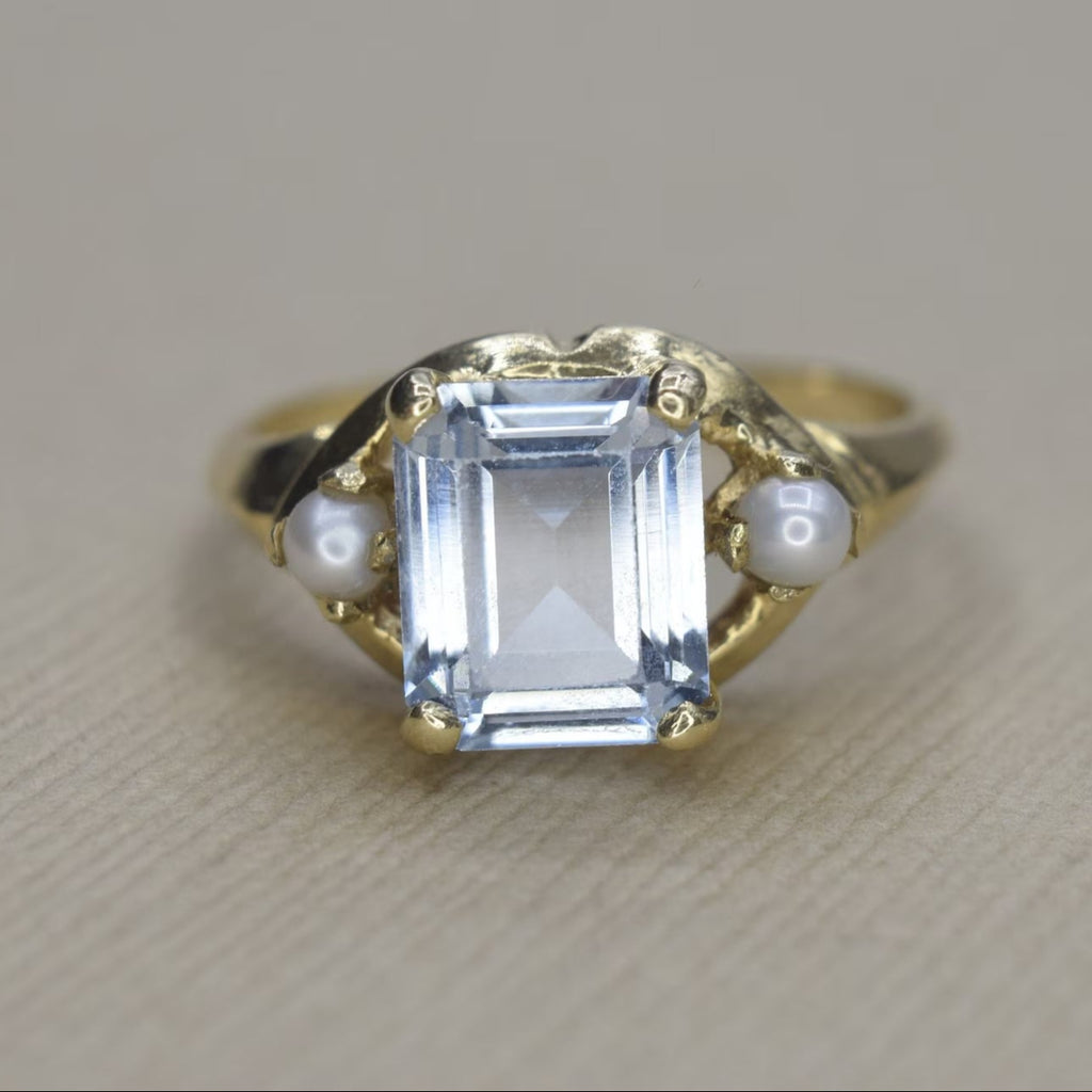 A vintage 14k gold ring with a rectangular cut synthetic blue spinel and seed pearl on each shoulder, displayed against a neutral background.