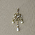 An antique Edwardian 14k gold diamond and baroque pearl lavaliere necklace with a single central diamond and five baroque pearls, displayed against a beige background.