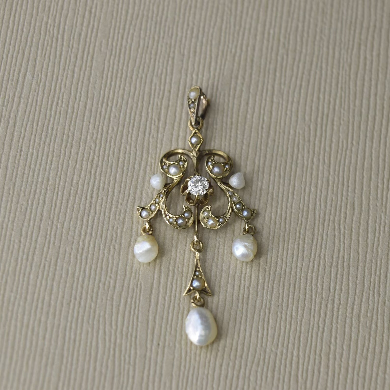 An antique Edwardian 14k gold diamond and baroque pearl lavaliere necklace with a single central diamond and five baroque pearls, displayed against a beige background.